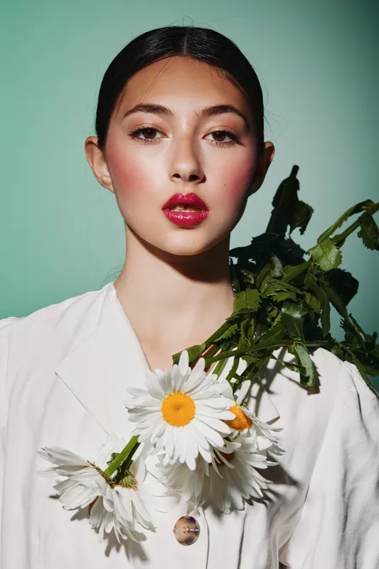 Young woman with glossy red lips and natural makeup holds fresh daisies wearing oversized white shirt. Clean mint background highlights beauty and fashion. Concept for beauty and creative fashion.