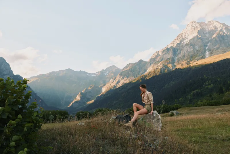 Woman lifestyle traveler sitting on rock in mountain valley, enjoying peaceful nature, sunny day, casual outfit, relaxed mood, scenic landscape background