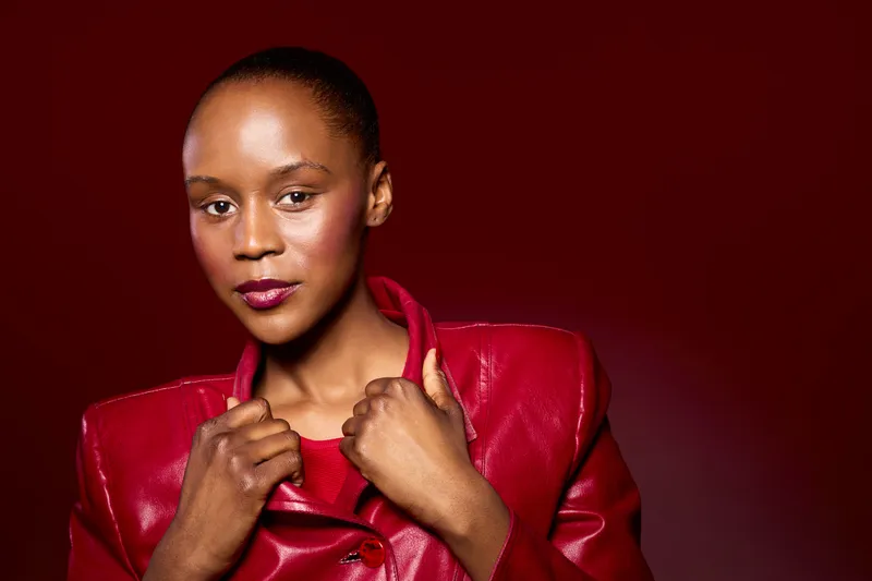 Confident African woman with dark skin wearing a red leather jacket poses against a deep red background. Portrait of elegance, fashion, and beauty with strong lighting highlights.