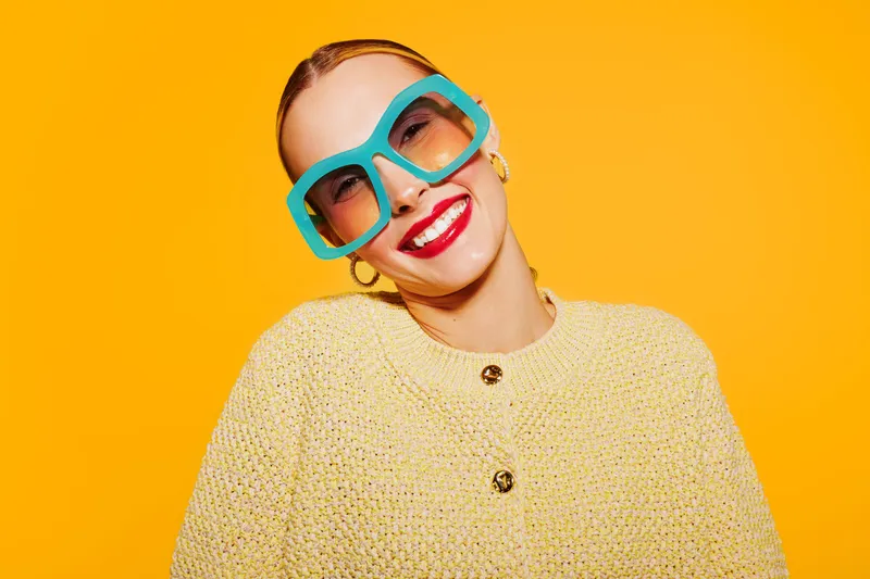 Smiling woman with large blue sunglasses and red lipstick wears yellow knitted sweater against bright orange background. Concept of fashion, style, confidence, and vibrant colors.
