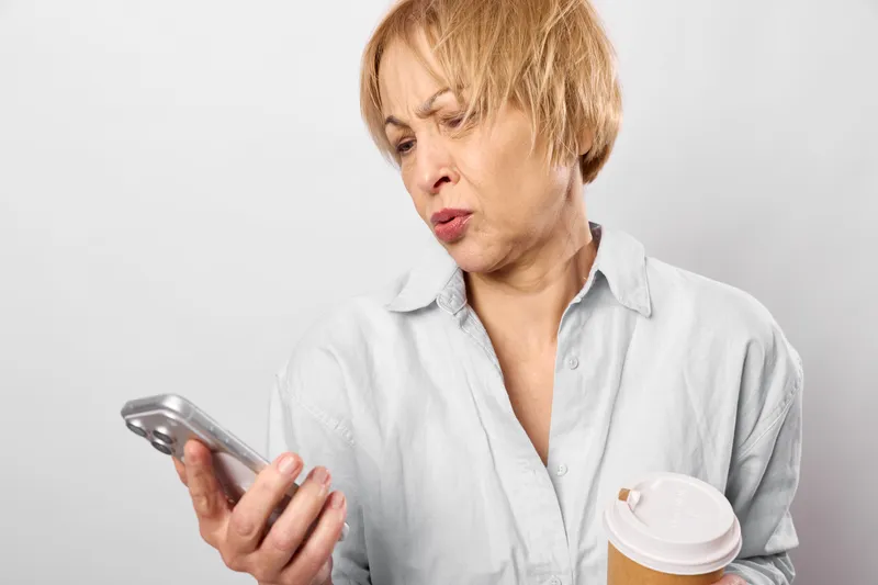 Woman with smartphone and cup looks worried, showing stress and health illness concern, indoors closeup portrait, holding takeaway beverage, neutral background.