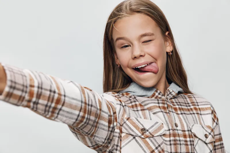 Teenage girl with long hair wearing plaid shirt making a playful face and sticking out tongue while taking a selfie on a light background.