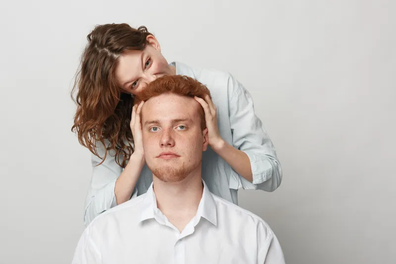 Young woman with brown hair gently massages head of serious redhead man in white shirts on light background. Concept of care, support, and intimate relationship between couple.