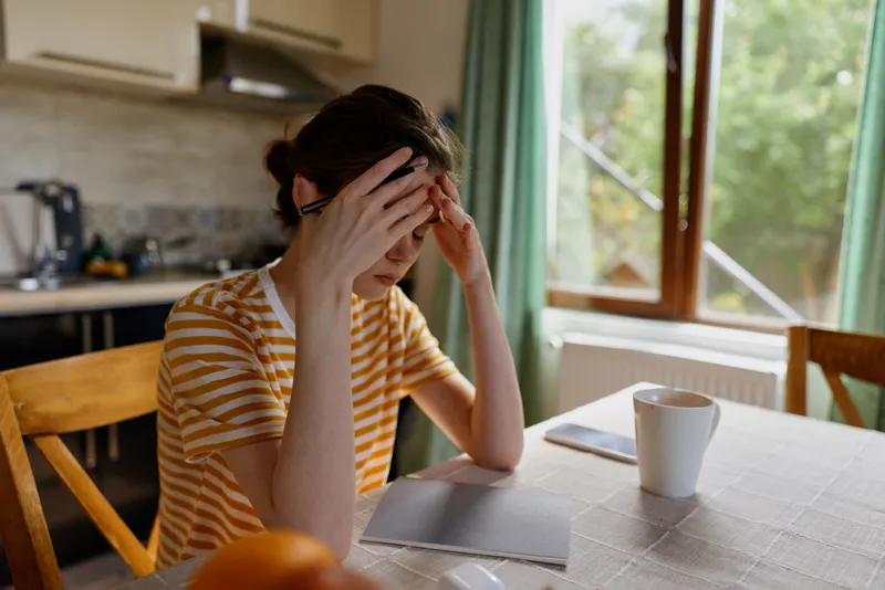 Stressed woman with striped shirt sits at a kitchen table, hands on her forehead, light streaming through a window, casual home setting, conveying fatigue, personal moments today right.
