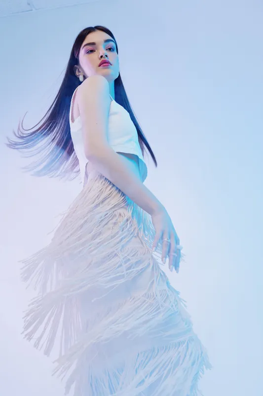 Young woman in white fringe dress posing with flowing hair against light background. Concept of beauty fashion elegance and style in studio lighting.