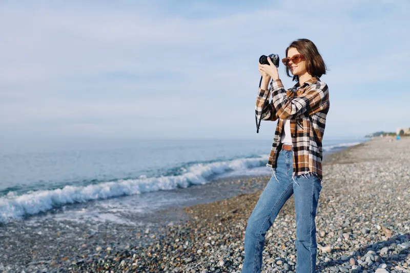 Photographer woman on a pebble beach filming with a camera, casual plaid shirt and jeans, ocean waves nearby, bright daylight capturing a creative outdoor moment