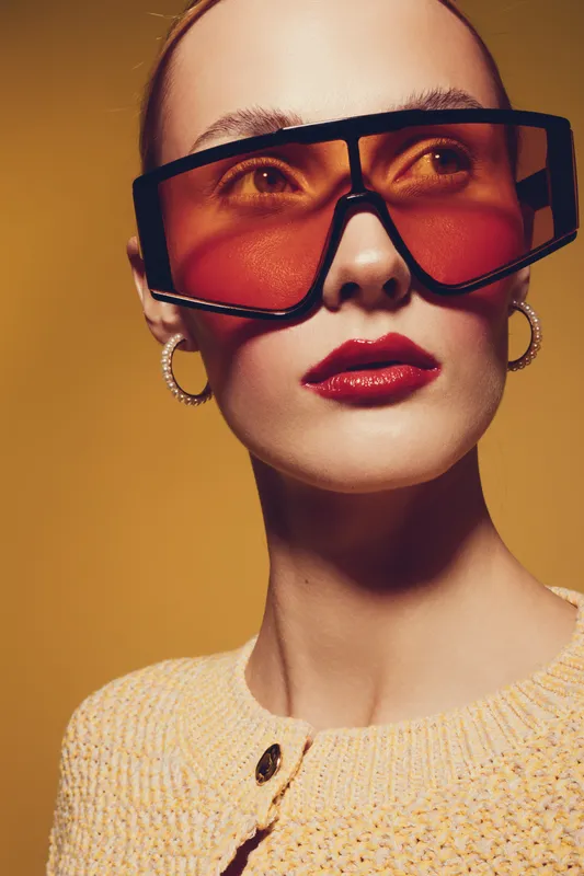 Close-up portrait of a stylish woman wearing oversized red sunglasses and gold hoop earrings. She wears a textured yellow sweater against a warm yellow background. Fashion and accessories concept.
