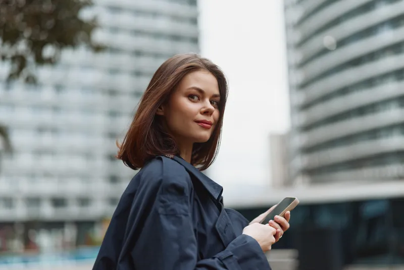 A young woman in a navy coat stands outdoors in a city setting, glancing back while holding a smartphone, conveying professional focus and urban energy.