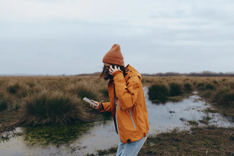 Woman lifestyle outdoors nature field walking hat orange coat, holding phone while walking on muddy path with puddles, cloudy sky above grass meadow, calm autumn mood