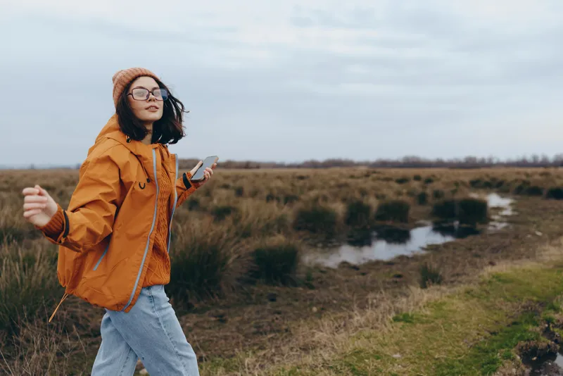 Woman outdoors in nature, orange jacket and sunglasses, walking near a river in an open landscape, lifestyle leisure with a relaxed smile on a spring day