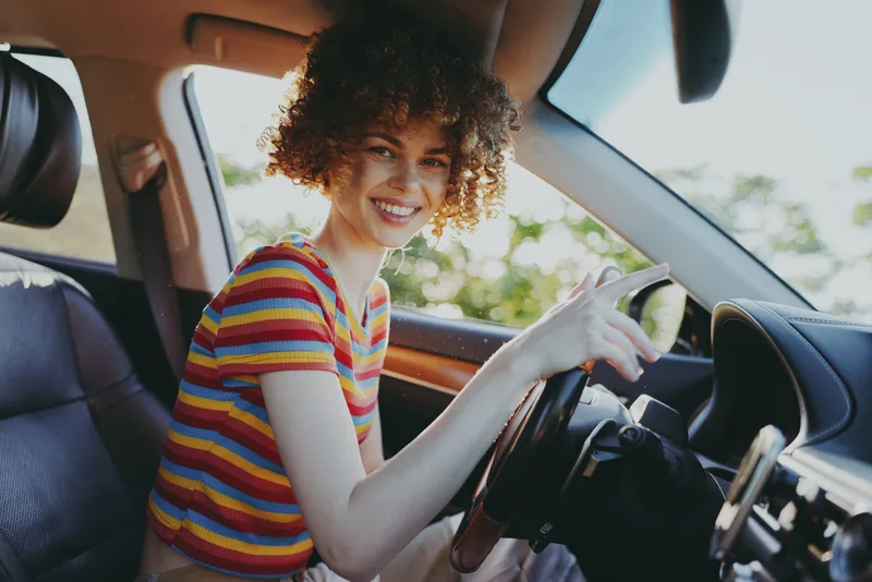 Cheerful woman driving, striped shirt, warm retro film color, candid smile, relaxed posture, sunlight through window, natural lighting, casual lifestyle, joyful moment inside car