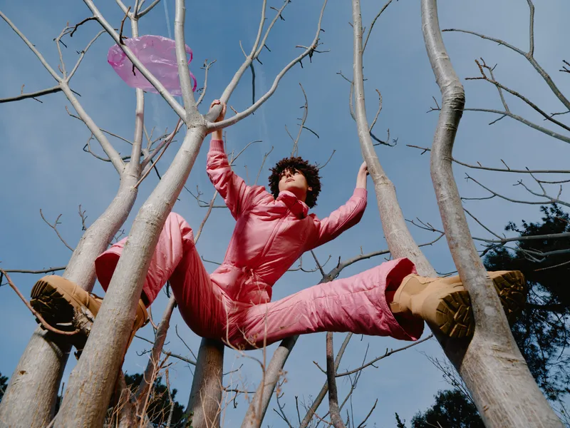 Person in pink jumpsuit climbs between tree branches, holding a pink frisbee, outdoors on a sunny day, playful athletic activity and carefree exploration