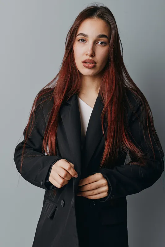 Young woman with long red hair wearing black blazer posing against gray background. Portrait of confident female model with natural makeup and serious expression in studio lighting.