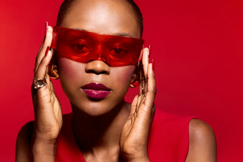 Portrait of a confident black woman with dark skin wearing red sunglasses and a red sleeveless top against a red background. Concept of fashion, style, beauty, and empowerment.