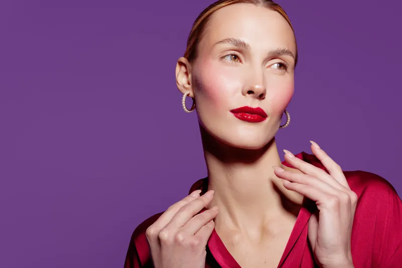 Portrait of a stylish woman with red lipstick and hoop earrings posing against purple background. Fashion concept with confident expression and elegant makeup in studio lighting.