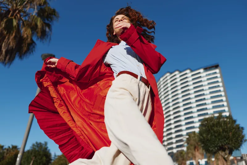 Confident woman outdoors wearing red coat and white pants walking with flowing coat in sunny urban setting with clear blue sky and modern building background