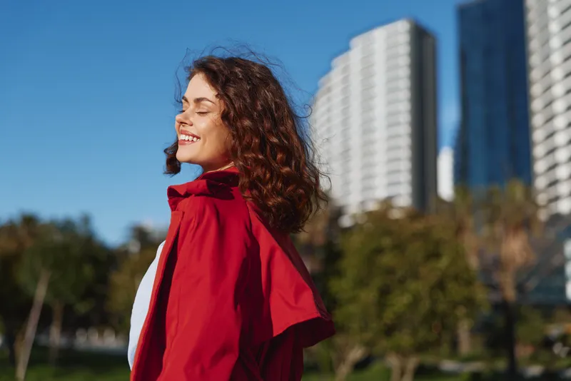 Cheerful woman outdoors, smiling in a red coat on a sunny day, relaxed pose, gentle breeze, urban park backdrop, vivid colors, natural light, greenery, warmth