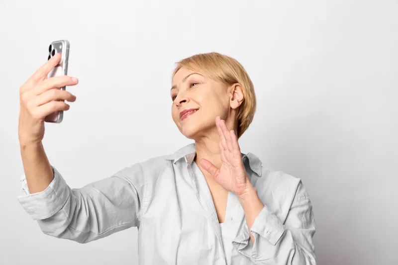 skincare, woman, elderly, selfie, smartphone, face, cream, cheek, close, studio lighting, smiling senior applies cream to cheek while viewing phone on white background
