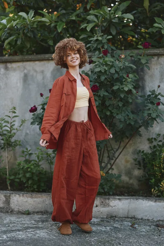 Woman lifestyle portrait outdoors in rust jacket and pants, cream top, curly hair, smiling confidently among greenery, relaxed fashionable vibe, casual elegant mood on sunny day, fresh atmosphere
