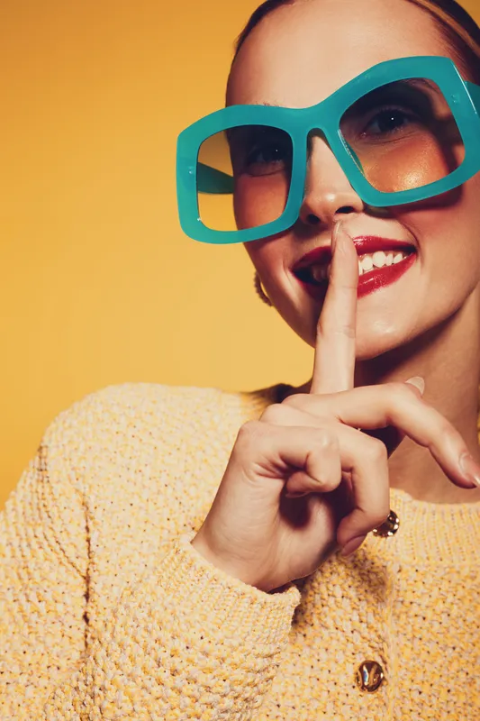 Smiling woman in large blue glasses and yellow sweater makes shushing gesture with finger on lips. Bright studio portrait with warm yellow background. Concept of secrecy and playful mood.
