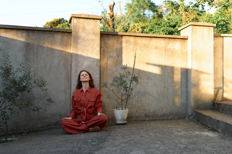 Calm woman sits cross legged in a red outfit on a concrete yard with a small potted plant, meditating pose, tranquil outdoor lifestyle moment, everyday relaxation scene