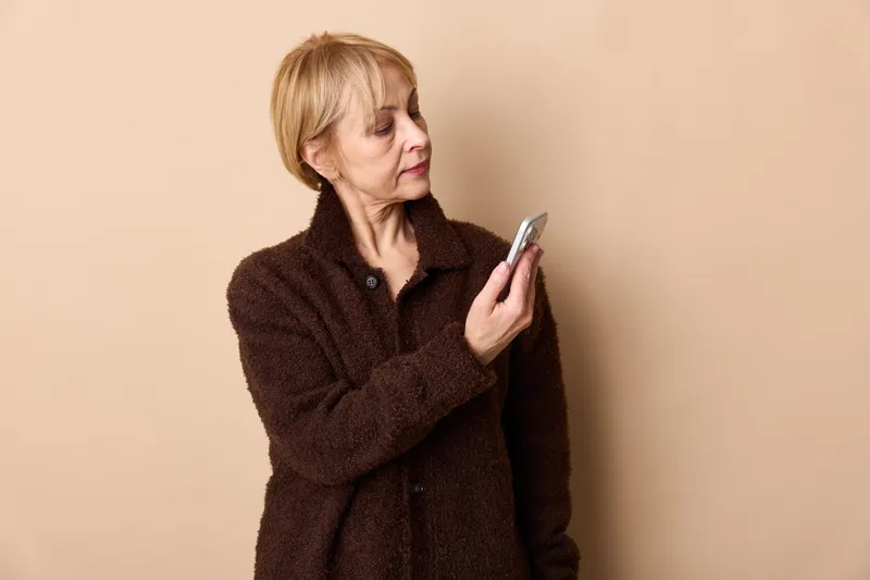 Elderly woman smartphone upset stressed loneliness sadness emotion mental health concept indoors portrait, woman in brown coat holding phone tense expression