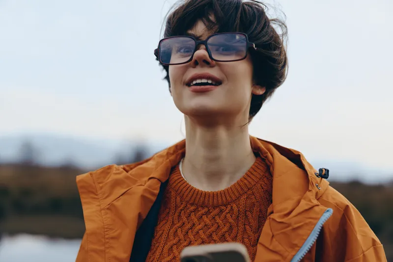 Woman outdoors wearing glasses and orange coat with backpack, with smile in winter air, close portrait with soft daylight, lifestyle concept of travel and casual style for women daily