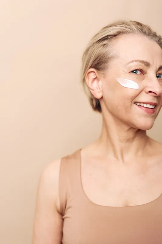 Smiling mature woman with short blonde hair applies white face cream on cheek. Close-up on beige background. Concept of skincare, aging skin care, moisturizing and natural beauty routine.