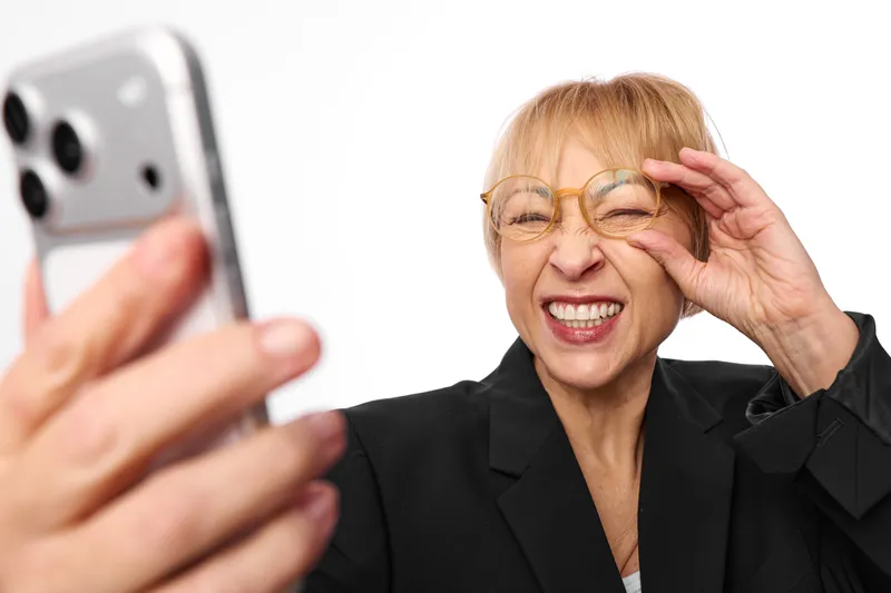 Elderly woman in business suit smiles and laughs while holding smartphone nearby, wearing eyeglasses, covering one eye with hand, indoor portrait with soft light