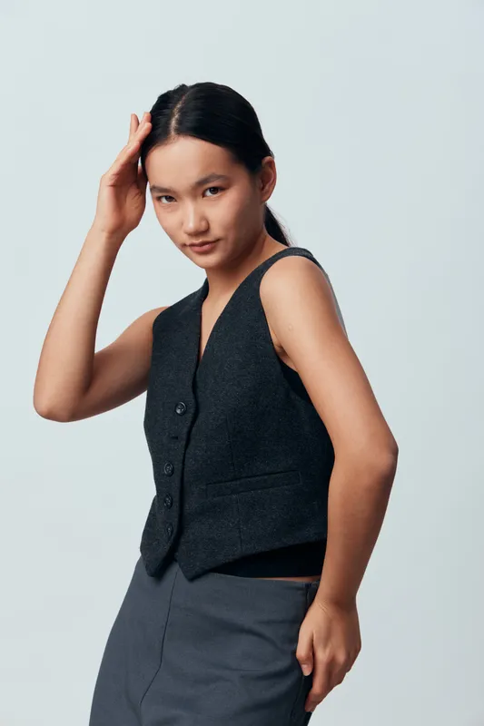 Young woman posing with confident expression in casual sleeveless vest and skirt against neutral background. Fashion model with natural look and hand near face in studio photo.