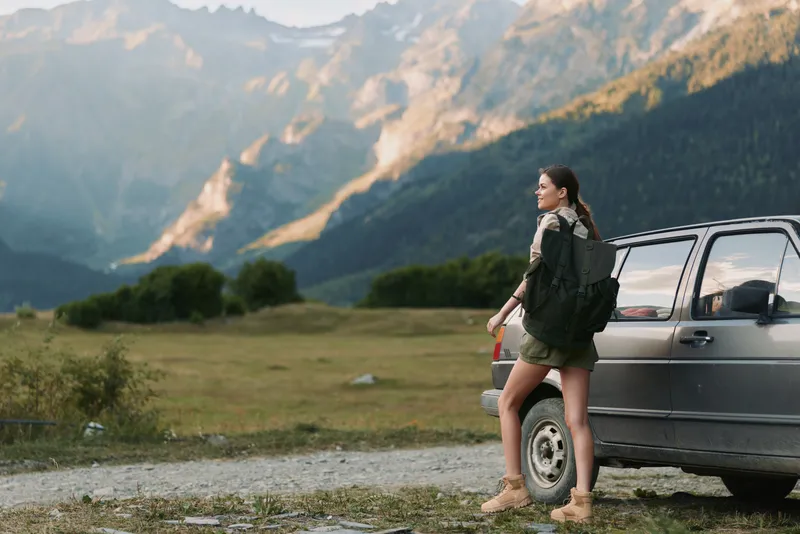 A young woman with a backpack stands beside a parked car in a rugged mountain landscape, looking toward the distant peaks, ready for an outdoor adventure today.