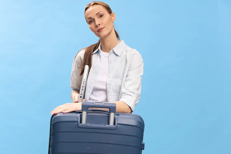 Young woman with flute and blue suitcase stands against blue background. Casual travel concept with musical instrument and relaxed mood. Studio shot with soft lighting.