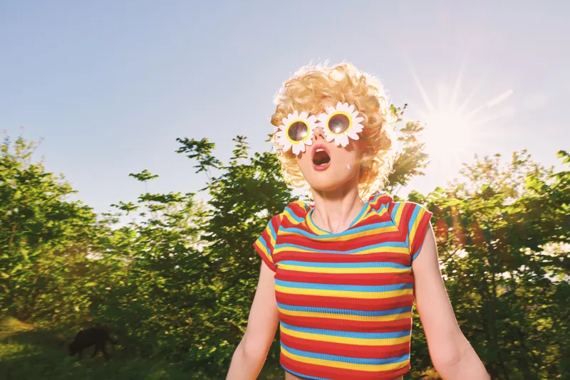 Cheerful woman outdoors, wearing a colorful striped top and flower sunglasses, laughing with head tilted, sunlit moment in a green park, relaxed lifestyle, retro mood and joyful energy fills the