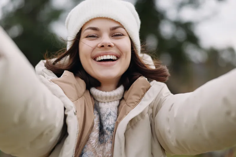 Joyful woman outdoors in winter attire, smiling broadly while taking a selfie in a cozy coat and beanie, capturing a sunny moment of happiness and fresh outdoor air.