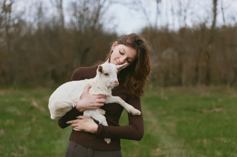 Young woman in brown sweater gently holds a white baby goat outdoors in a green field during early spring, showing a tender connection with the animal and a calm natural mood.