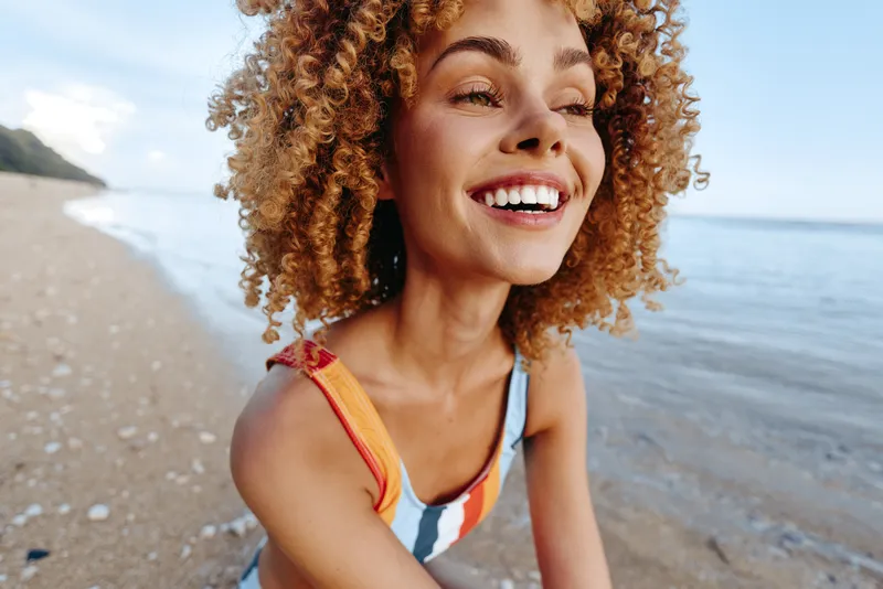 Young woman with curly hair smiles joyfully while sitting on a sandy beach near the calm sea under a bright sky on a warm day.