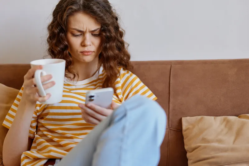 A stressed woman sits on a brown sofa at home, holding a mug in one hand and scrolling on her phone. A candid lifestyle moment showing fatigue, distraction, and everyday domestic life.