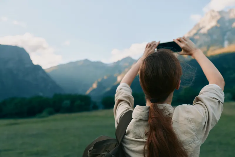 Cheerful woman outdoors, capturing a mountain valley scene with a smartphone, casual jacket, backpack, and relaxed posture, sunlit greens and fresh breeze, lifestyle moment