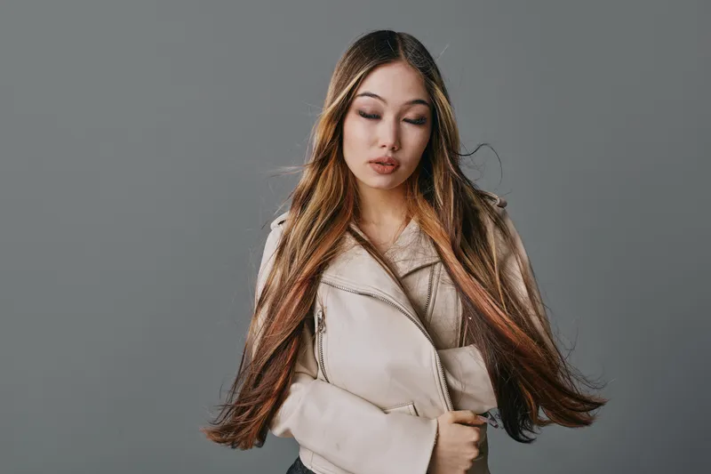 Young woman with long straight hair and neutral makeup wearing beige jacket standing with eyes closed on gray background. Beauty, fashion, portrait and style concept with casual clothing.
