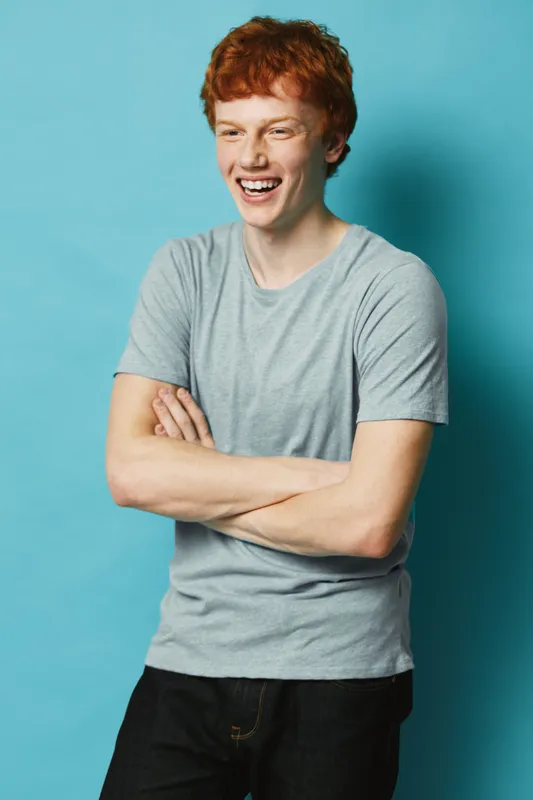 Young man with red hair laughing and standing with arms crossed against blue background. Casual outfit with gray t-shirt and dark pants in studio portrait.