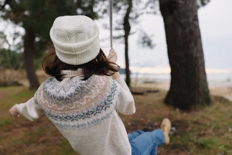 Back view of a person on a swing in a park, wearing a knit sweater and beanie, enjoying a calm outdoor moment among trees with soft autumn light today.