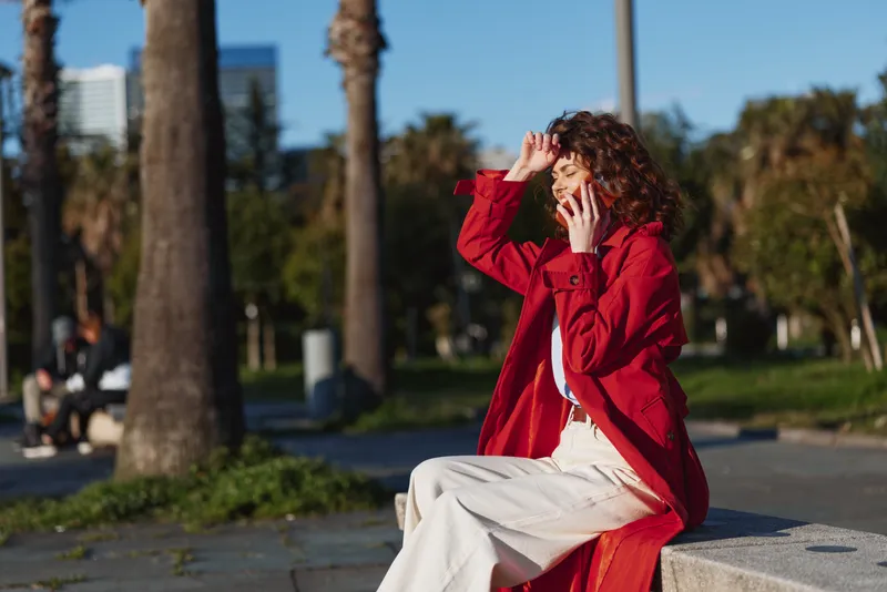 One woman with curly hair in a red coat sits on a concrete ledge in a sunny park, adjusting her hat and looking away, creating a candid urban portrait.