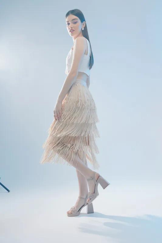 Young woman in a stylish beige fringe skirt and white top poses elegantly against a soft light blue background. Concept of woman beauty fashion and modern style.