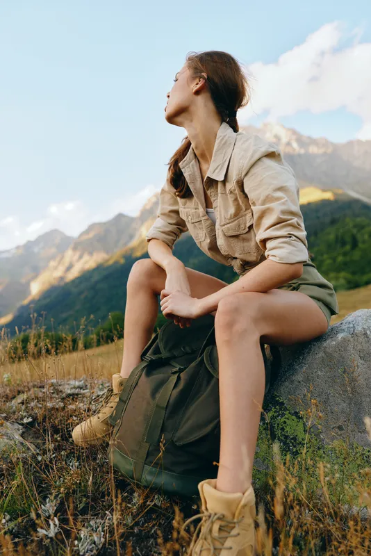 Young woman rests on a rock with a backpack in a mountain landscape, conveying calm exploration and outdoor adventure, with soft sunlight