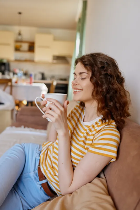 A woman relaxing at home, sipping coffee from a mug in a bright living space. A casual, cozy lifestyle moment showcasing everyday comfort and calm in a domestic setting.