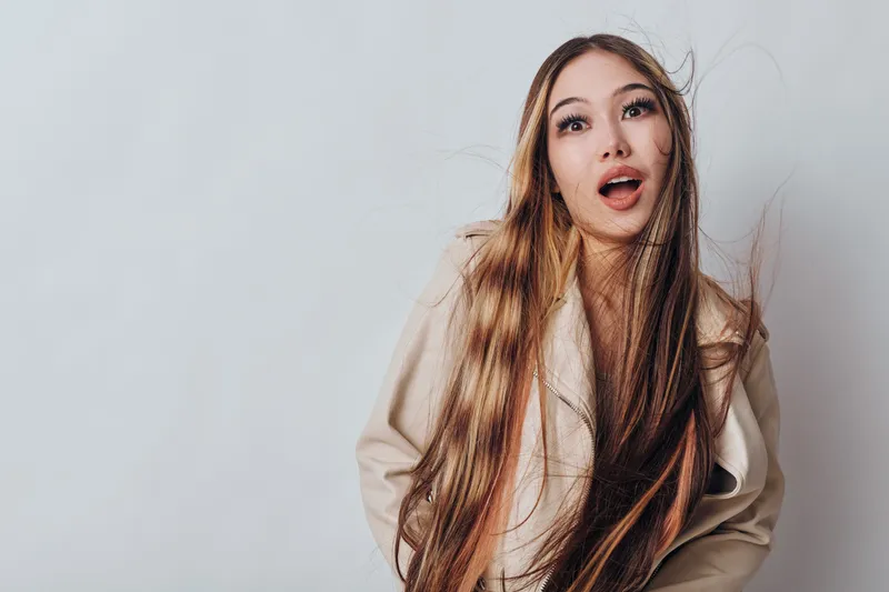 Young woman with long hair and surprised expression wearing beige jacket with open mouth and wide eyes on white background, casual portrait with natural light, beauty and emotion.