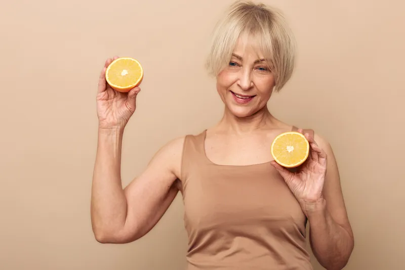 Smiling mature woman with short blonde hair holds two orange halves in hands against beige background. Concept for healthy lifestyle, nutrition, wellness, and natural vitamin intake.