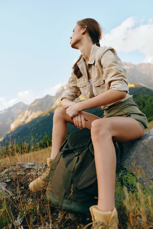 Young woman rests on a rock during a mountain hike, gazing at rugged peaks with a calm, focused expression.