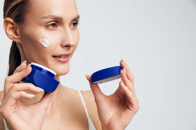 Young woman applying white face cream on cheek holding blue jar and lid. Skincare routine concept with natural light and soft focus on neutral background.