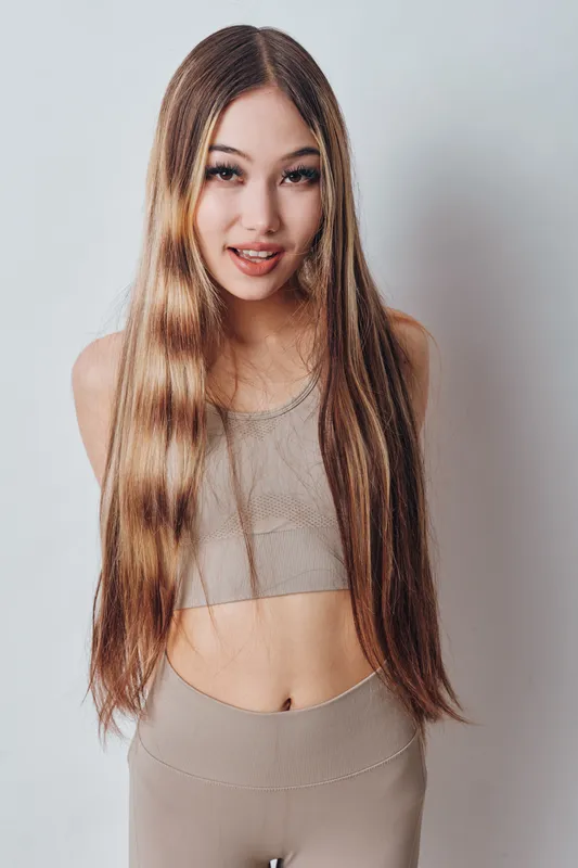 Young woman with long straight hair wearing beige knitted top and high-waisted pants posing smiling against light background in studio portrait.